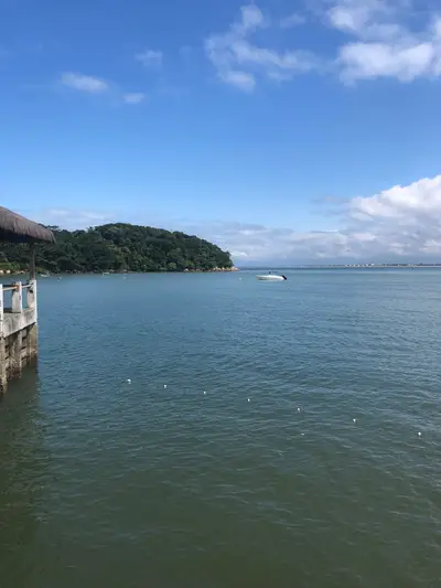 Vista de águas calmas junto a um trapiche, com barco ancorado e morro coberto pela mata ao fundo. A luz diurna intensa realça o azul do céu e os reflexos na superfície da baía.