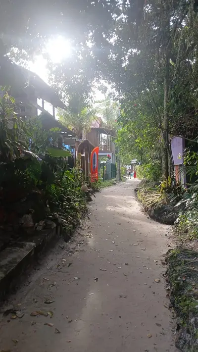 Trilha de areia entre construções rústicas e vegetação densa, com pessoas ao fundo. A luz solar atravessa a copa das árvores, criando um ambiente tropical e acolhedor.