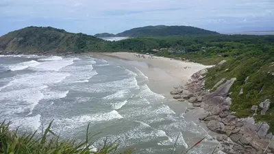 Vista elevada de uma praia de areia clara, com ondas quebrando junto a costões rochosos e mata densa ao redor; pequenas pessoas aparecem espalhadas pela faixa de areia. A cena tem luz diurna suave, sob céu parcialmente nublado, destacando o contraste