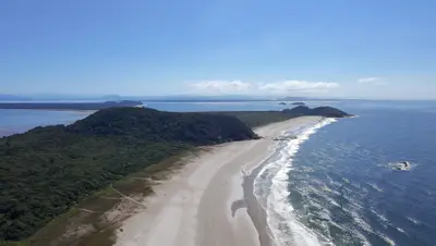 Vista aérea de uma praia extensa, com faixa de areia clara, mar azul e vegetação rasteira que avança até a mata. Uma trilha corta o terreno em direção à costa, sob luz solar intensa e céu limpo.