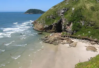 Praia de areia clara entre costões rochosos e mata verde, com pequenas ondas e algumas pessoas no mar. Um passarela de madeira acompanha as pedras sob luz solar intensa e céu aberto.