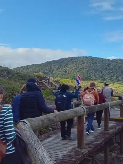 Pessoas percorrem uma passarela de madeira em meio à vegetação, diante de um morro coberto por mata. O ambiente é de trilha ao ar livre, sob luz natural suave e céu azul com nuvens.