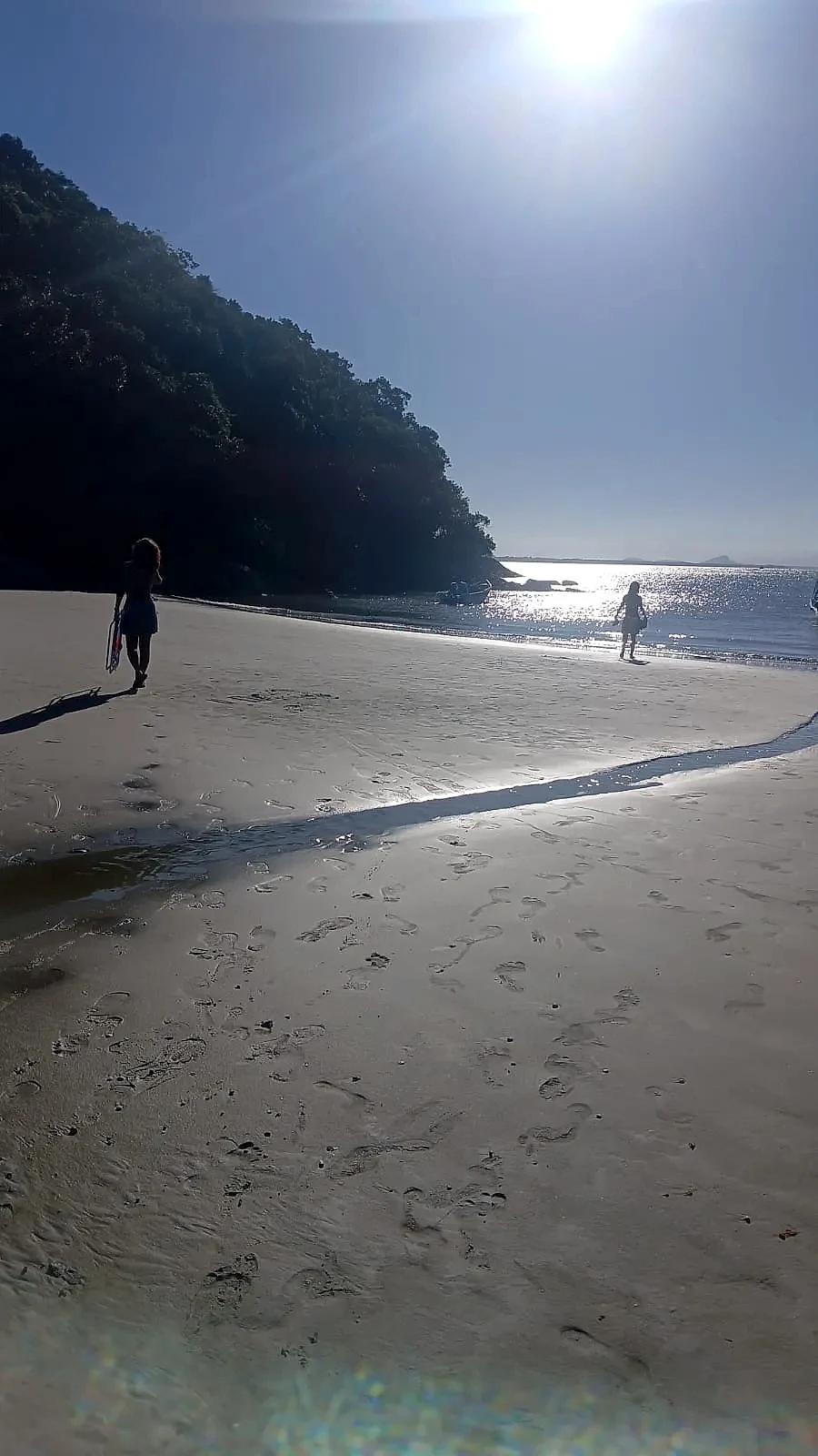 Praia de areia clara junto à mata, com duas pessoas caminhando perto da água e marcas de passos na faixa de areia. A luz forte do sol cria reflexos intensos no mar e sombras longas, em um ambiente tranquilo.