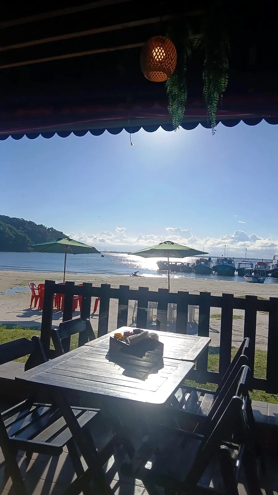 Varanda de restaurante com mesas de madeira voltada para a praia, onde guarda-sóis e barcos estão ancorados junto à água calma. A luz intensa do sol reflete no mar sob céu azul, criando um ambiente litorâneo acolhedor.