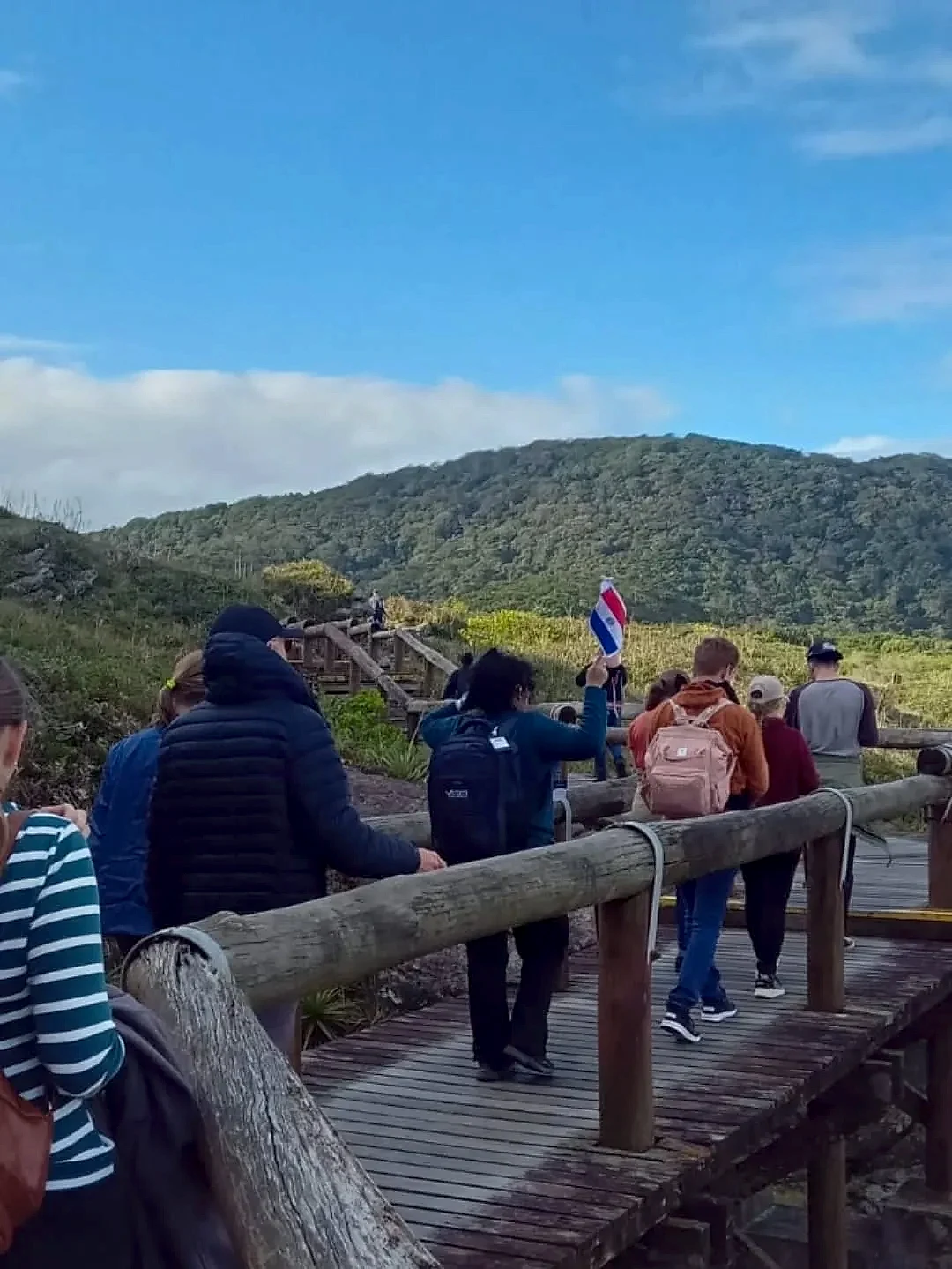 Pessoas percorrem uma passarela de madeira em meio à vegetação, diante de um morro coberto por mata. O ambiente é de trilha ao ar livre, sob luz natural suave e céu azul com nuvens.
